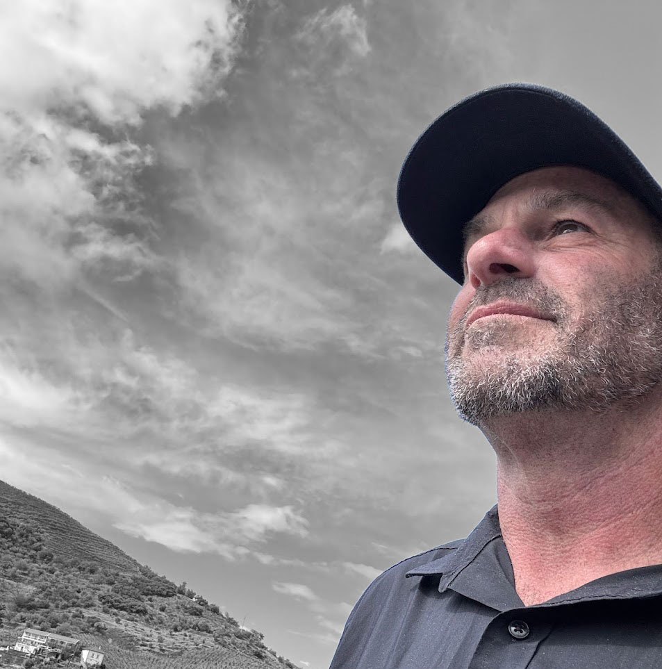 Rich Cohen in black and white, wearing a cap and looking up toward a cloudy sky