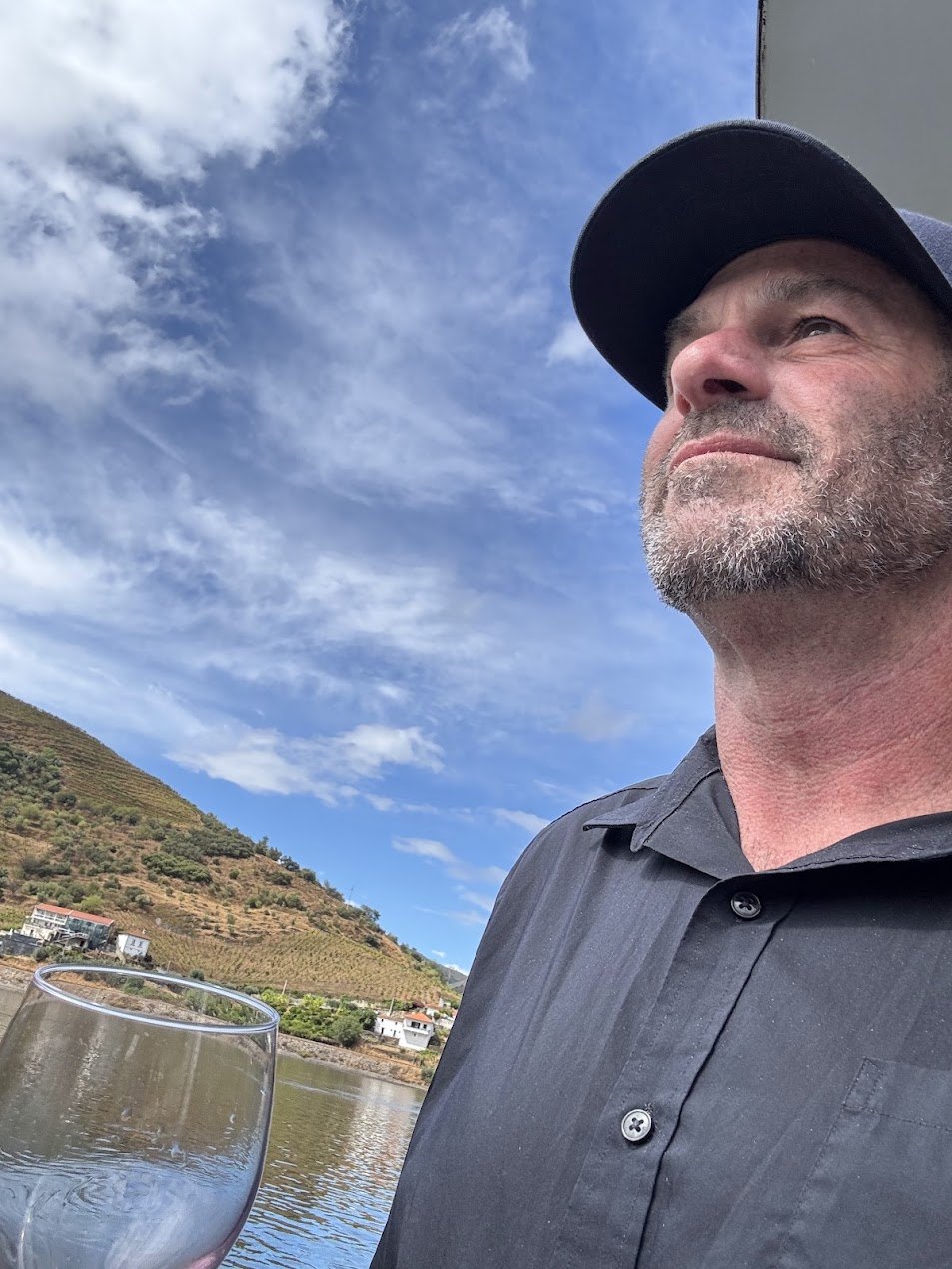 Rich Cohen in a dark cap looking up at a blue sky, with a river and terraced vineyard hills behind him and a wine glass in the foreground